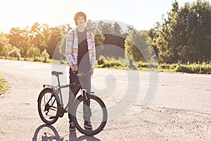 Healthy lifestyle concept. Teenage male with trendy hairstyle wearing casual shirt, jeans and sport shoes standing with bicycle on