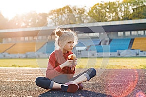 Healthy lifestyle and healthy food concept. Little beautiful girl child in sportswear eating apple sitting on stadium after