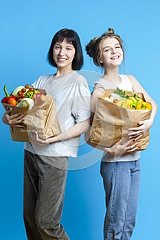Healthy Lifestyle Ideas. Two Smiling Caucasian Girlfriends Posing With Paper Bag Filled Grocery And Vegetables Posing Over Pure