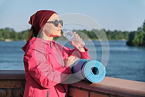 Healthy lifestyle of mature woman, outdoor portrait of an age female in sportswear with yoga mat, drinking water from bottle