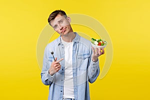 Healthy lifestyle, people and food concept. Reluctant handsome young man pointing finger at disgusting salad, unwilling
