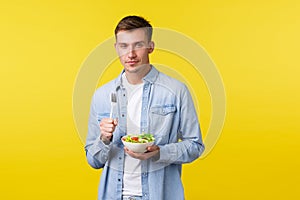 Healthy lifestyle, people and food concept. Sassy good-looking blond man eating salad, squinting and smiling delighted