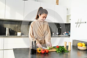 Healthy lifestyle. Young woman in bathrobe preparing food, chopping vegetables, cooking dinner on kitchen counter
