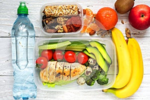 Healthy lunch boxes with chicken, fresh vegetables, fruits and nuts with bottle of water on white wooden background