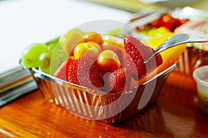 Healthy man using a fork to eat in the office a delicious fresh fruit salad on aluminium box on wooden table