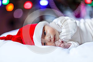 Healthy newborn baby in Santa hat near Christmas tree with colorful garland lights on background. Closeup of cute child