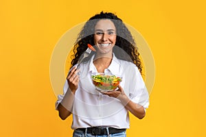 Healthy Nutrition. Cheerful Young Woman Eating Vegetable Salad, Standing Over Yellow Background