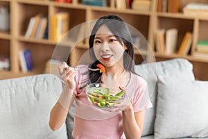 Healthy nutrition concept. Young asian lady eating yummy vegetable salad on sofa at home, smiling at camera