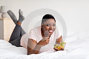 Healthy nutrition for weight control. Curvy black woman eating delicious vegetable salad, lying on bed at home