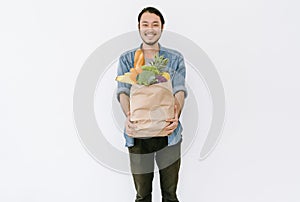 Healthy positive young Asian man holding paper shopping bag full of fresh vegetables and groceries with copy space isolated on