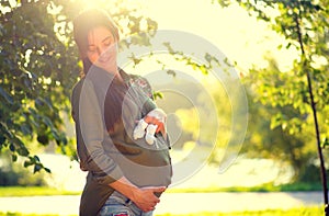 Healthy pregnancy concept. Pregnant woman touching her belly outdoor, holding and playing with baby shoes