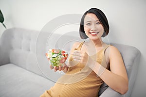 Healthy pregnancy food. Asian girl eating fresh vegetable salad, resting on sofa at home