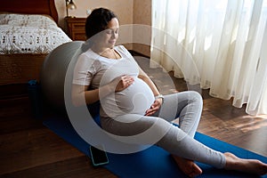 Healthy pregnancy and maternity concept. Portrait of a sporty pregnant gently stroking her belly, sitting on yoga mat