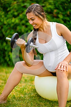Healthy Pregnant Woman Lifting Dumbbells On Exercise Ball In Park