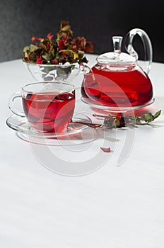 Healthy red herbal tea in transparent teapot, cup with hawthorn berries, rose hip, dry leaves in sunlight in contrast black.