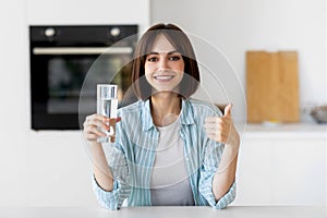 Healthy refreshment. Happy young lady drinking water from glass and showing thumb up, sitting in kitchen