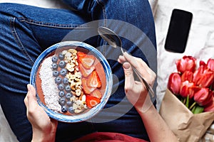 A healthy romantic breakfast: a bowl of smoothies in woman` s hands in jeans, a bouquet of flowers and a phone on the