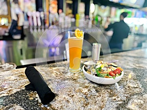 Healthy salad bowl, black rolled cloth napkins, tall pilsner beer glass with orange slide cut and blurry bartender serving beer