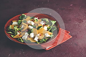 Healthy salad with persimmon, doucette lambs-lettuce, cornsalad, feld salad and feta cheese on a red plate on a red background.