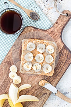 Healthy sandwich with crunchy peanut butter, banana and chia seeds, on wooden board with cup of coffee, top view, vertical