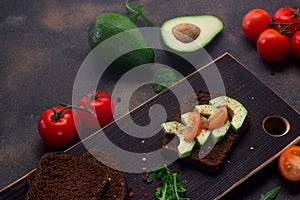 Healthy sandwich on a wooden cutting board with avocado, tomatoes and spices on whole grain bread. Dark background