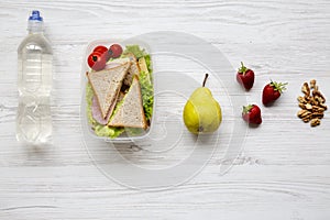 Healthy school lunch box with fresh organic vegetables sandwich, walnuts, fruits and bottle of water on white wooden background, f