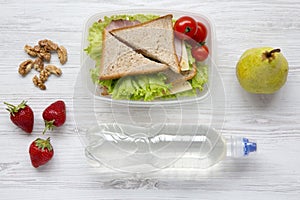 Healthy school lunch box with fresh organic vegetables sandwiches, walnuts, bottle of water and fruits on white wooden background,