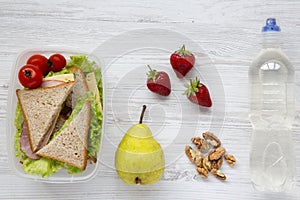 Healthy school lunch box with fresh organic vegetables sandwiches, walnuts, bottle of water and fruits on white wooden background,