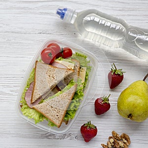 Healthy school lunch box with fresh organic vegetables sandwiches, walnuts, bottle of water and fruits on white wooden surface, o