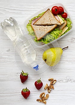 Healthy school lunch box with fresh organic vegetables sandwiches, walnuts, bottle of water and fruits on a white wooden table,