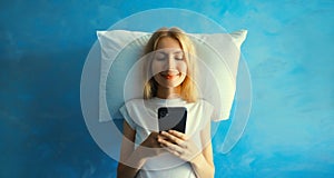 Healthy sleep, happy relaxed sleepy young woman with mobile phone on white soft comfortable pillow imagining she lying on the bed