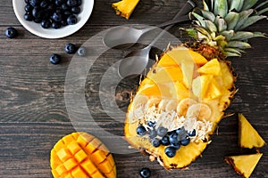 Smoothie bowl in a pineapple with coconut, bananas, mango & blueberries, top view on dark wood