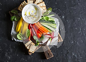 Healthy snack - raw vegetables and yogurt sauce on a wooden cutting board, on a dark background, top view. Vegetarian healthy food photo