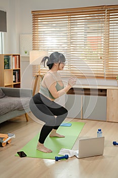 A healthy, strong Asian woman in sportswear is doing a squad on a yoga mat, working out at home