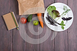 Healthy vegetarian food concept - paper eco bag with healthy various vegetables, plate with greenery and blank notepad for recipe