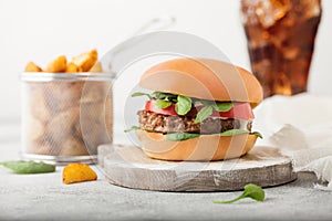 Healthy vegetarian meat free burger on round chopping board with vegetables on light table background with potato wedges and glass