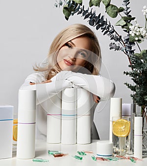 Healthy wealthy blonde woman in white stylish pullover sits at table leaning on set of jars and tubes of cosmetic means