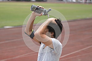 Healthy young Asian runner pouring water with waterbottle on his face after running on track in stadium.