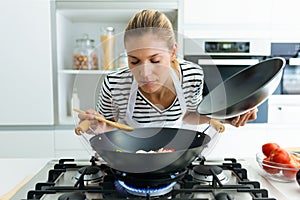 Healthy young woman cooking and smelling food in frying pan in the kitchen at home.