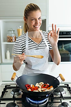 Healthy young woman looking at camera while cooking and mixing food in frying pan in the kitchen at home.