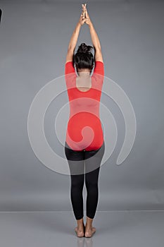 Healthy young woman in red t-shirt and black leggings doing yoga and stretching exercises isolated on grey background