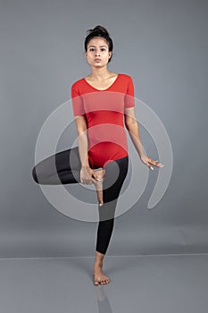 Healthy young woman in red t-shirt and black leggings doing yoga and stretching exercises isolated on grey background