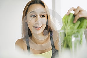 Healthy young woman in sport bra at kitchen making fresh vegetable juice in juicer blending machine, preparing nutritious vitamin