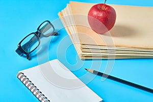 Heap of blank magazines, newspapers or some documents and red apple, notepad, pencil, glasses on blue desk