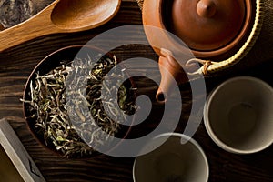 Heap of dried, raw white tea leaves in wooden bowl with teapot and cups on wooden table flat lay top view from above