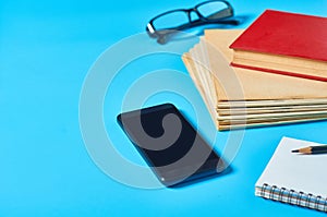 Heap of magazines, newspapers or some documents and red apple, notepad, pencil, glasses, smartphone on blue desk
