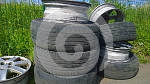Heap of old used car tires and aluminum rims on a ground against green grass background. Concept of problem of recycling tyre and