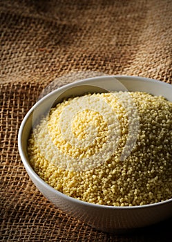 Heap of raw, uncooked couscous in white bowl on burlap