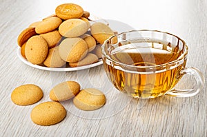 Heap of round biscuits in plate, transparent cup with tea, few cookies on wooden table