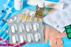 Heap of white pills and yellow capsules, bottle, syringes, bandage, patches and ampoules with red vaccine on blue background